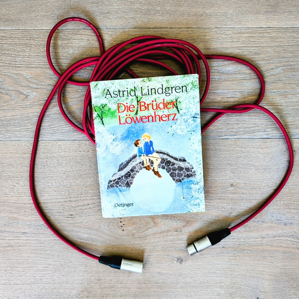 Das Bild zeigt das Buch DIE BRÜDER LÖWENHERZ von Astrid Lindgren; es liegt auf einem roten Mikrofon-Kabel.