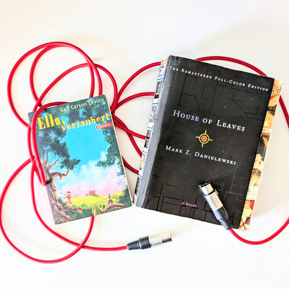Das Foto zeigt links das Buch ELLA, VERZAUBERT von Gail Carson Levine und rechts daneben die englische Originalausgabe des Buchs DAS HAUS. HOUSE OF LEAVES von Mark Z. Danielewski, das in der Übersetzung von Christa Schuenke bei Klett-Cotta erschienen ist. Die Bücher liegen auf einem roten Mikrofonkabel.

