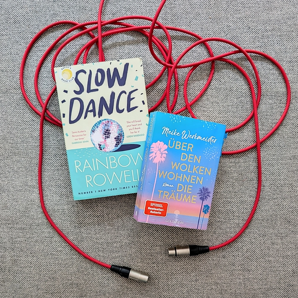 Das Foto zeigt links den Roman SLOW DANCE von Rainbow Rowell und rechts daneben den Roman ÜBER DEN WOLKEN WARTET DAS GLÜCK von Meike Werkmeister; beide Bücher liegen auf einem roten Mikrofonkabel.