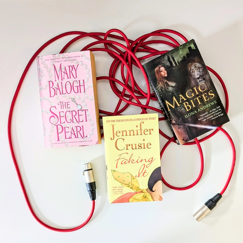 Auf dem Bild sind drei Bücher zu sehen, über die Anne Rudolph im Buchpodcast RABABUMM spricht: THE SECRET PEARL von Mary Balogh, FAKING IT von Jennifer Cruside und MAGIC BITES von Ilona Andrews. Die drei Bücher liegen auf einem roten Mikrofonkabel.