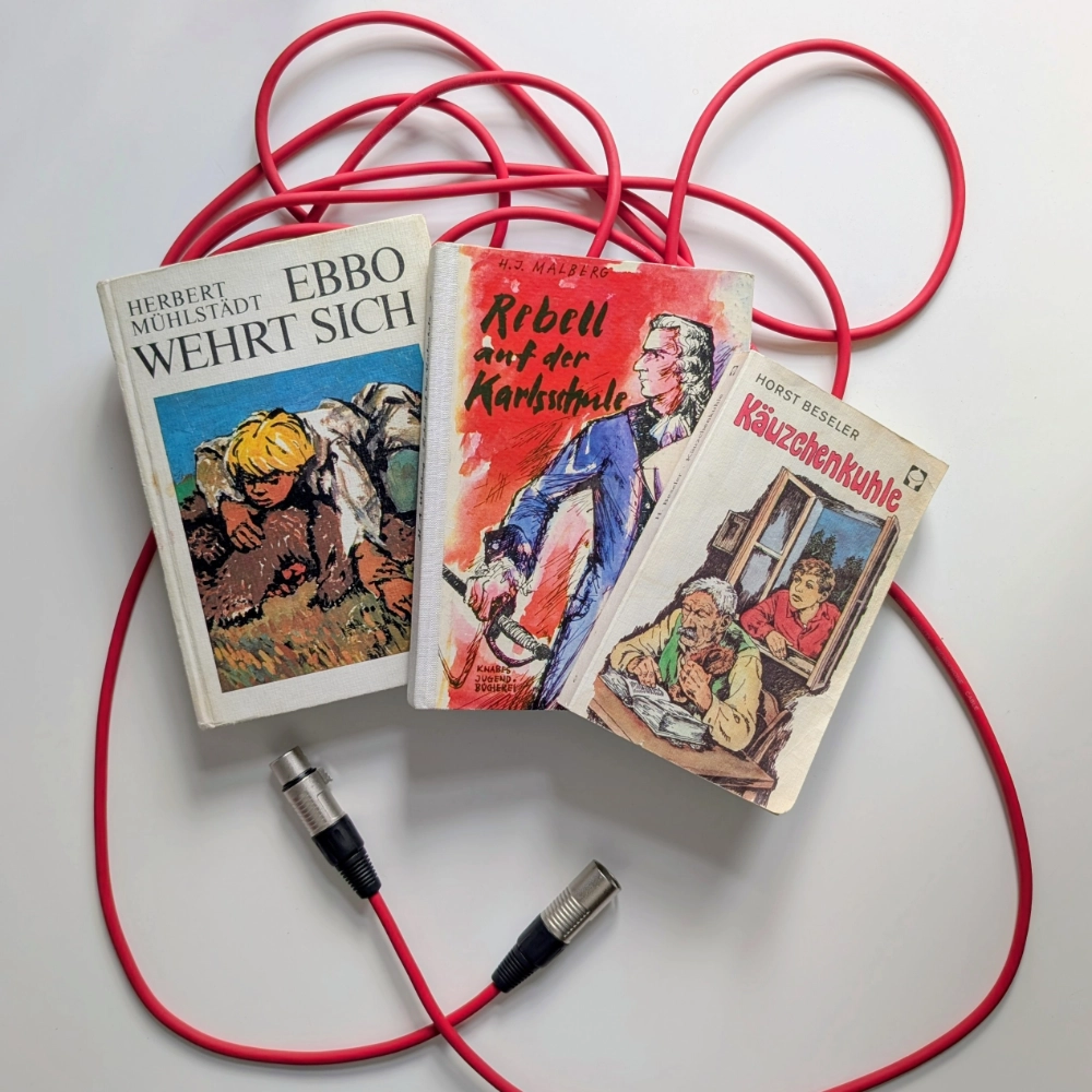 Das Foto zeigt drei Kinder- und Jugendromane, die auf einem roten Mikrofonkabel liegen: EBBO WEHRT SICH von Herbert Mühlstädt, REBELL AUF DER KARLSSCHULE von H.J. Malberg und KÄUZCHENKUHLE von Horst Beseler.
