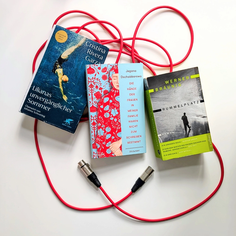 Das Foto zeigt die drei Romane LILIANAS UNVERGÄNGLICHER SOMMER von Cristina Rivera Garza, DIE HÄNDE DER FRAUEN IN MEINER FAMILIE WAREN NICHT ZUM SCHREIBEN BESTIMMT von Jegana Dschabbarowa und RUMMELPLATZ von Werner Bräuning. Die Bücher liegen auf einem roten Mikrofonkabel.