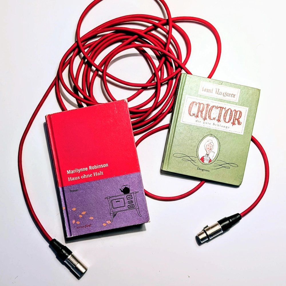 Das Foto zeigt die beiden Bücher CRICTOR – DIE GUTE SCHLANGE von Tomi Ungerer, erschienen bei Diogenes, und HAUS OHNE HALT von Marilynne Robinson, in der Übersetzung von Karen Nölle erschienen beim Verlag Edition fünf. Beide Bücher liegen auf einem roten Mikrofonkabel.