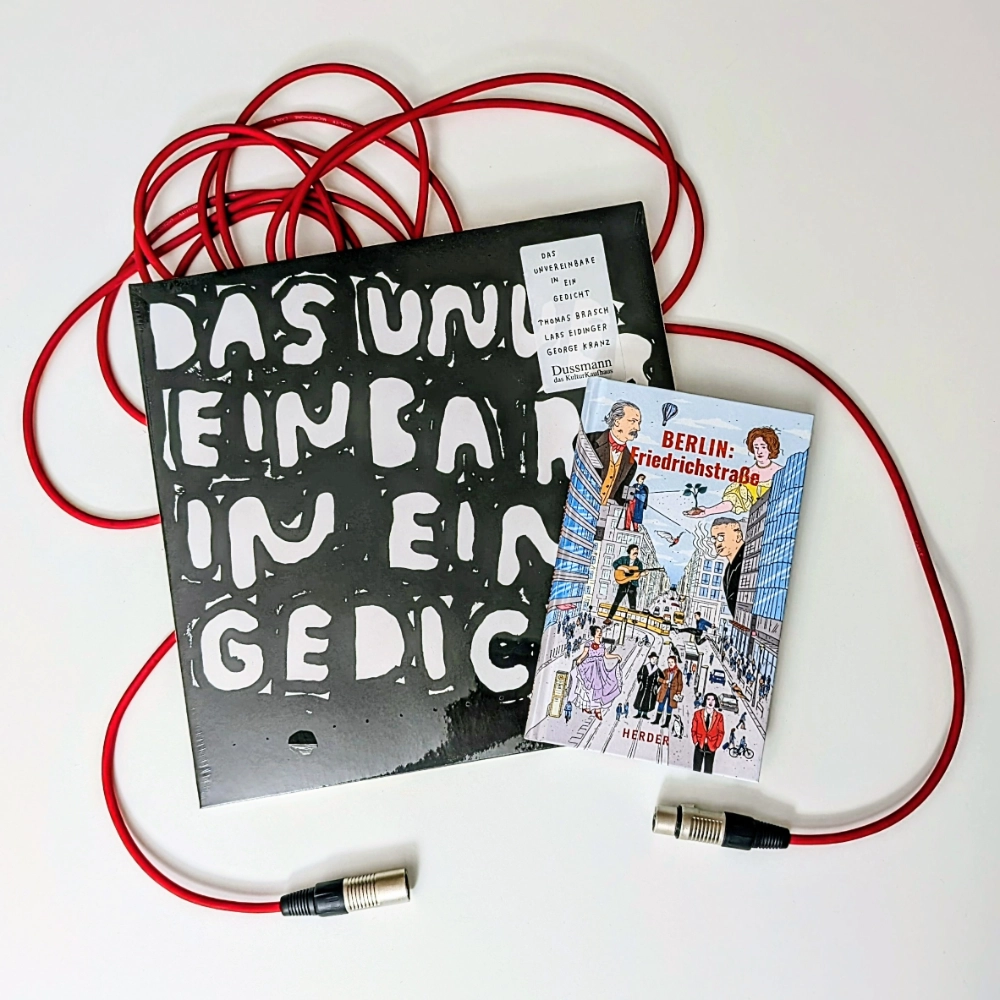 Das Foto zeigt die Schallplatte DAS UNVEREINBARE IST EIN GEDICHT und das Buch BERLIN: FRIEDRICHSTRASSE, die beide exklusiv bei Dussmann - Das Kulturkaufhaus erhältlich sind; beides liegt auf einem roten Mikrofonkabel.