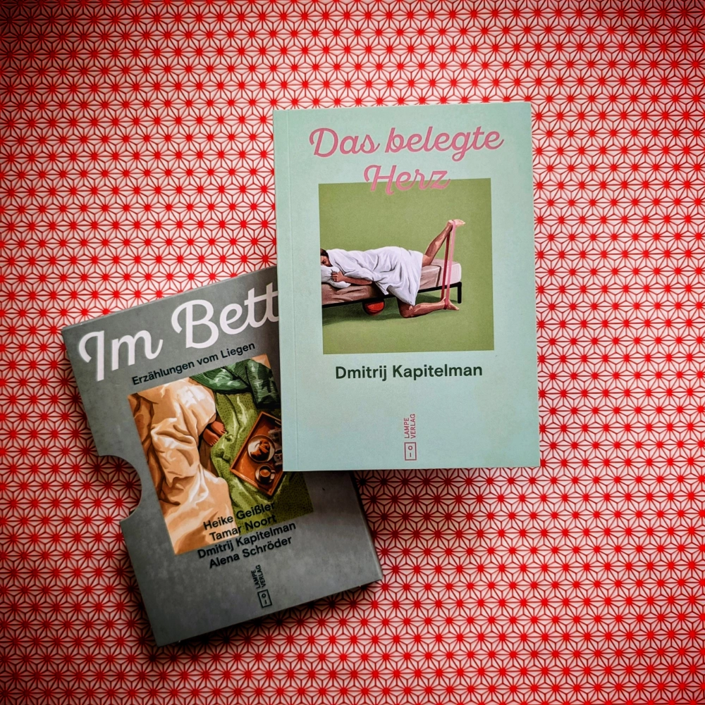 Das Foto zeigt das Minibuch „Das belegte Herz“ von Dmitrij Kapitelmann. Es ist Teil des Buchschubers „Im Bett – Erzählungen vom Liegen“, erschienen im Lampe Verlag. Das Minibuch und der Buchschuber liegen auf einem gemusterten Papierhintergrund.