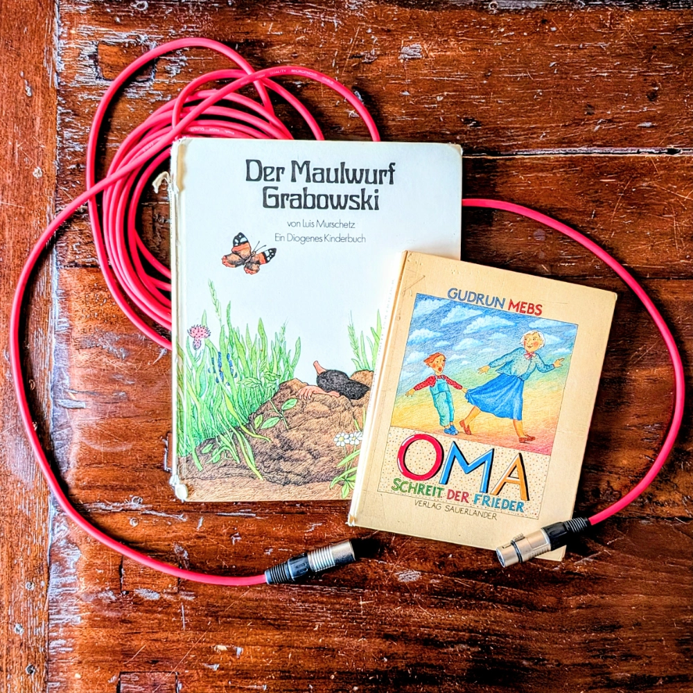 Das Foto zeigt die beiden Bücher DER MAULWURF GRABOWSKI und OMA, SCHREIT DER FRIEDER. Die beiden Bücher liegen auf einem roten Mikrofonkabel und einem Holztisch.