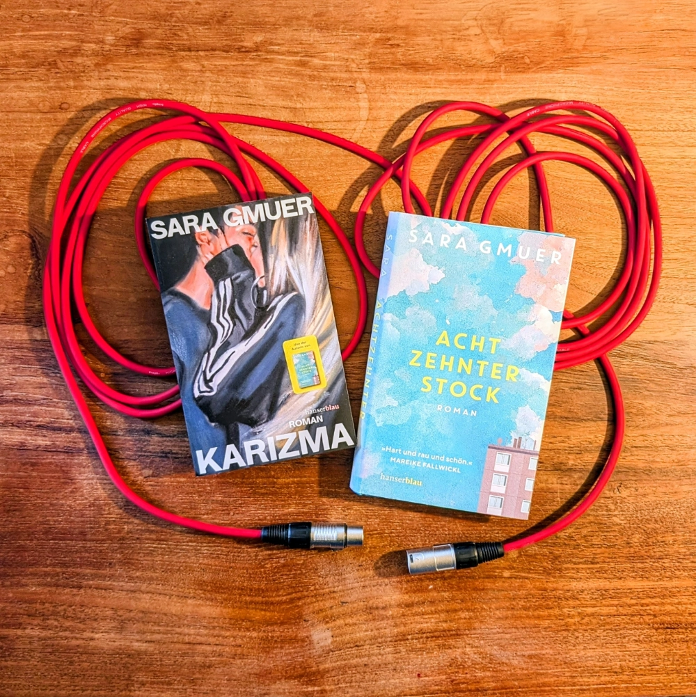 Das Foto zeigt die Romane KARIZMA und ACHTZEHNTER STOCK von Sara Gmuer. Die Bücher liegen auf einem roten Mikrofonkabel auf einem Holztisch.