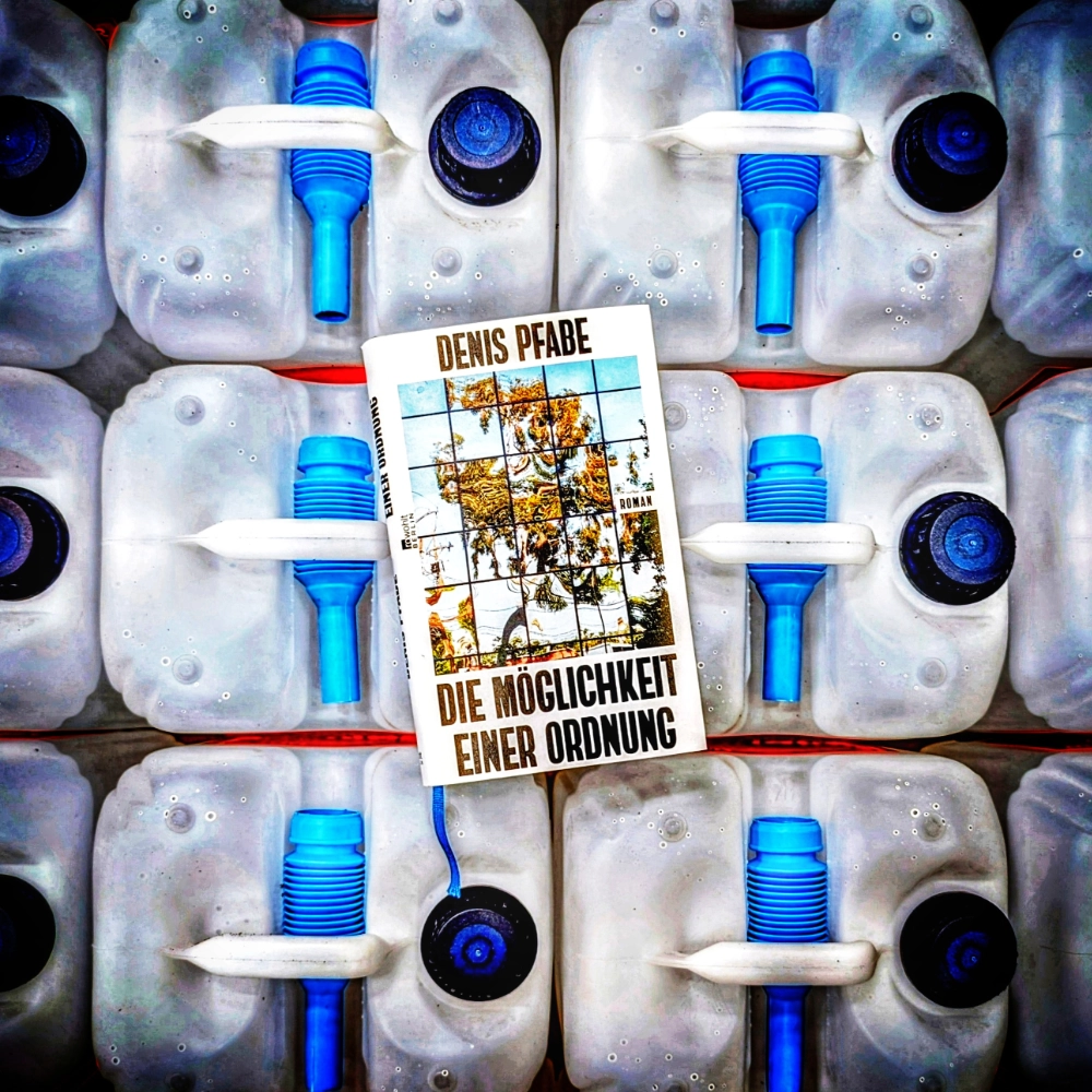 Das Foto zeigt die Hardcover-Ausgabe des Romans „Die Möglichkeit einer Ordnung“ von Denis Pfabe, der bei Rowohlt Berlin veröffentlicht wurde. Das Buch liegt auf Kanistern mit Kühlflüssigkeit.