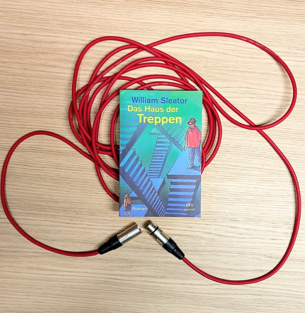 Das Bild zeigt das Buch DAS HAUS DER TREPPEN von William Sleater; es liegt auf einem roten Mikrofonkabel.
