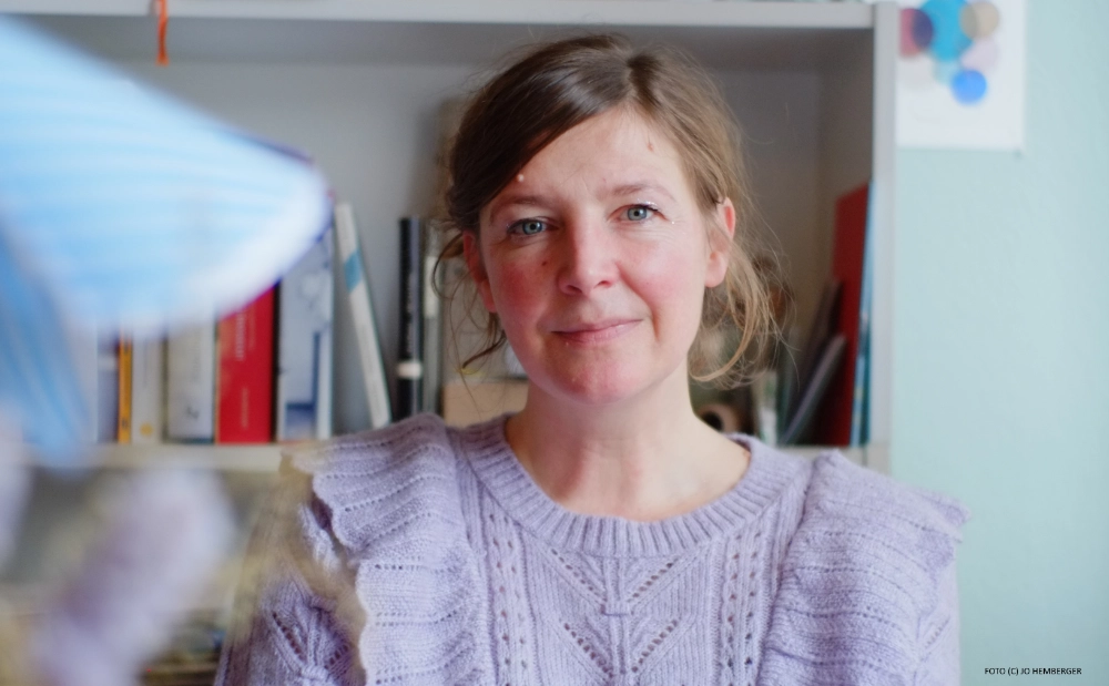 Das Foto von Jo Hemberger zeigt die Autorin Simone Scharbert, die an ihrem Arbeitsplatz vor einem Buchregal sitzt.