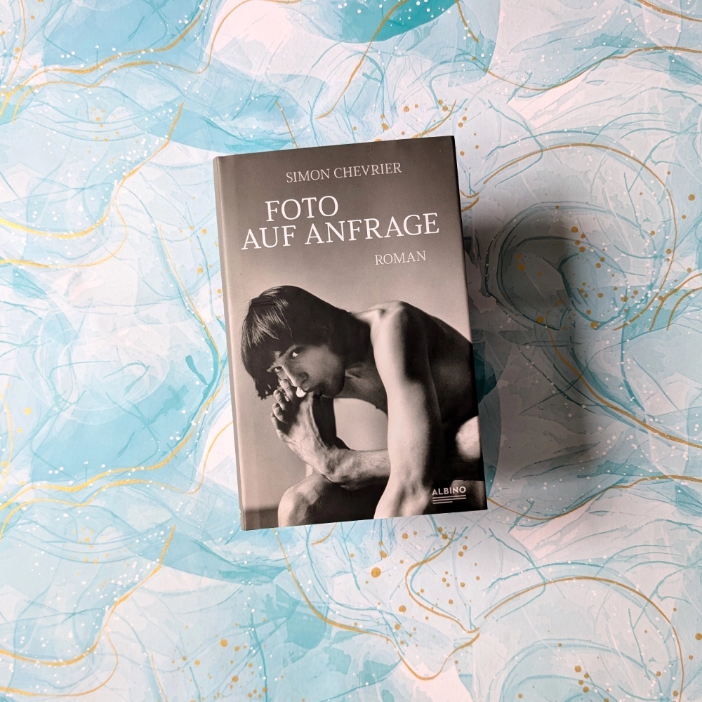 Das Foto zeigt die Hardcover-Ausgabe des Romans „Foto auf Anfrage“ von Simon Chevalier, der im Albino Verlag veröffentlicht wurde. Das Buch liegt auf einem gemusterten Papier-Hintergrund.
