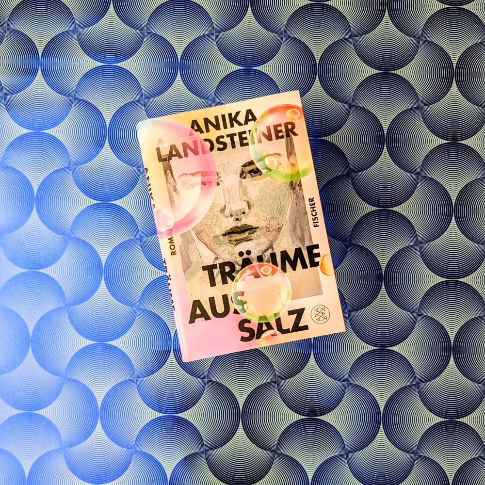 Das Foto zeigt den Roman "Träume aus Salz" von Anika Landsteiner; das Buch liegt auf einem gemusterten Papierhintergrund.